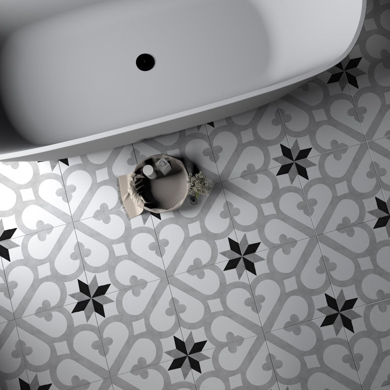 May include: A white bathtub with a black drain sits on a floor covered in square tiles featuring a repeating pattern of gray heart-like shapes and black eight-pointed stars. A small round basket containing white items sits on the tiled floor.