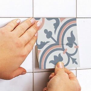 Puede incluir: Un adhesivo cuadrado para azulejos con un patr&oacute;n decorativo en azul apagado y rosa suave sobre un fondo blanco. El adhesivo se est&aacute; aplicando sobre una superficie de azulejos blancos, mostrando un dise&ntilde;o sim&eacute;trico de inspiraci&oacute;n floral.