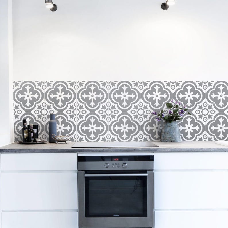May include: A kitchen backsplash featuring square tiles with a repeating grey and white quatrefoil pattern. The tiles are installed behind a stainless steel oven and a white countertop, with a grey vase containing purple flowers on the right.