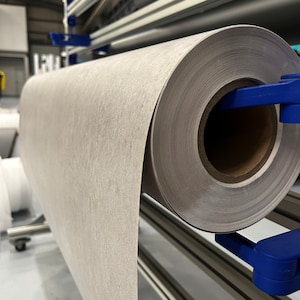 K&ouml;nnte beinhalten: Eine gro&szlig;e Rolle hellgrauen, strukturierten Materials wird von blauen Klammern gehalten. Das Material scheint Papier oder Stoff zu sein und ist auf einem Metallrahmen montiert. Die Rolle ist teilweise abgerollt und zeigt die Textur.