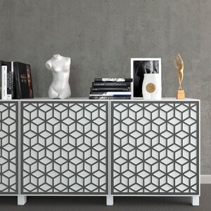 K&ouml;nnte beinhalten: Ein wei&szlig;er Schrank mit geometrisch gemusterten T&uuml;ren, der B&uuml;cher, eine B&uuml;stenskulptur, ein gerahmtes Bild, eine Vase und eine goldfarbene Figur enth&auml;lt. Der Schrank steht vor einer grauen Wand und schafft eine moderne &Auml;sthetik.