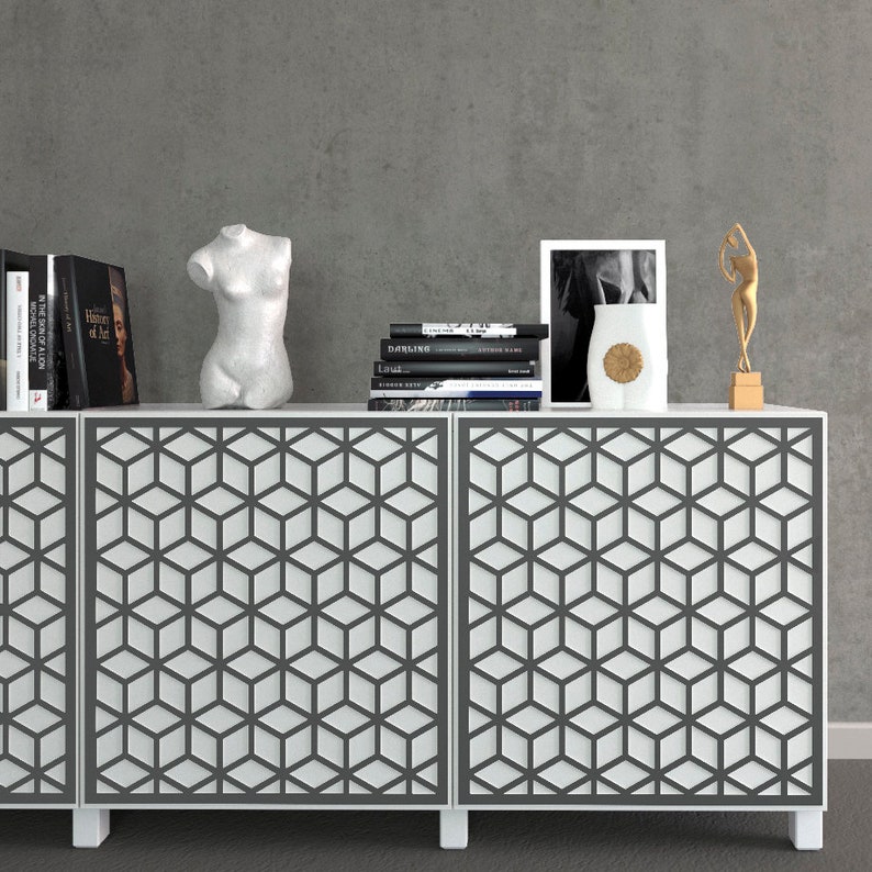K&ouml;nnte beinhalten: Ein wei&szlig;er Schrank mit geometrisch gemusterten T&uuml;ren, der B&uuml;cher, eine B&uuml;stenskulptur, ein gerahmtes Bild, eine Vase und eine goldfarbene Figur enth&auml;lt. Der Schrank steht vor einer grauen Wand und schafft eine moderne &Auml;sthetik.