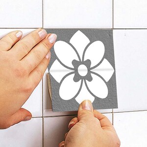 May include: A square tile sticker featuring a gray background with a white six-petaled flower design. The sticker is being applied to a white tiled surface, showing a textured finish on the gray portion and a smooth white floral pattern.