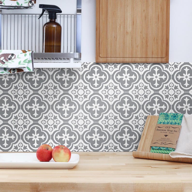 May include: A kitchen backsplash featuring square tiles with a repeating grey and white quatrefoil pattern. The wall is positioned above a light wood countertop with a white rectangular dish holding two apples and a beeswax wrap set.