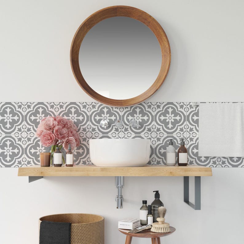 May include: A bathroom vanity area featuring a round wooden mirror above a white vessel sink on a light wood floating shelf. The wall has gray and white patterned tiles. Below, a woven basket and a small wooden stool hold various toiletries.