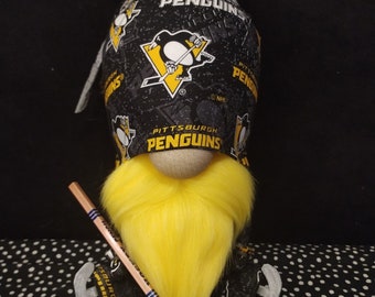 May include: A gnome doll wearing a black hat and outfit with the Pittsburgh Penguins logo and team name. The gnome has a yellow beard and is holding a hockey stick. The doll is sitting on a black and white polka dot surface.