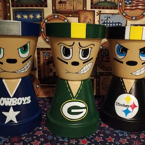 Puede incluir: Tres macetas de terracota pintadas con caras de dibujos animados y logotipos de equipos de la NFL. Las macetas están pintadas en los colores de los Dallas Cowboys, los Green Bay Packers y los Pittsburgh Steelers. Las macetas están decoradas con los logotipos de los equipos y las palabras "Cowboys", "Packers" y "Steelers".