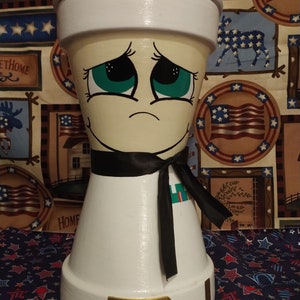 Peut inclure: Un pot en céramique blanc en forme de marin avec un ruban noir et une bordure dorée. Le pot a un visage peint avec des yeux verts et une expression triste.