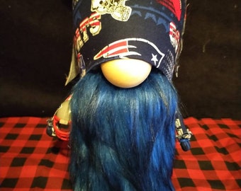 Pode incluir: Um gnomo azul e branco com uma longa barba azul. O gnomo está usando um chapéu com o logotipo do New England Patriots e as palavras "Patriots" e "D.N.M."