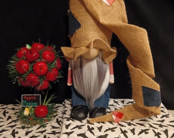 May include: A burlap gnome with a long, flowing burlap hat and a white beard. The gnome is wearing a red and white checked shirt and blue jeans. The gnome is standing on a white fabric with a black crow pattern. The gnome is holding a sign that says "Apple Picking".