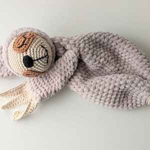 May include: A crocheted sloth toy with a wooden ring attached. The sloth is light pink with a brown face and black eyes. The toy is perfect for a baby or toddler.