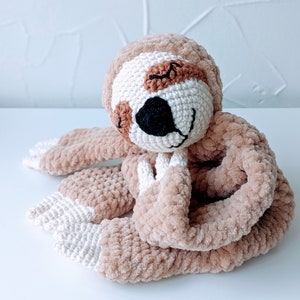 May include: A crocheted sloth toy with brown fur, white accents, and a black nose. The sloth is curled up in a sleeping position.