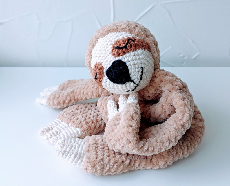 May include: A crocheted sloth toy with brown fur, white accents, and a black nose. The sloth is curled up in a sleeping position.
