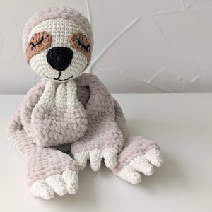 May include: A crocheted sloth toy with a light gray body, white feet, and brown eyes. The sloth is sitting with its hands clasped in front of it.