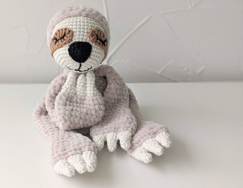 May include: A crocheted sloth toy with a light gray body, white feet, and brown eyes. The sloth is sitting with its hands clasped in front of it.