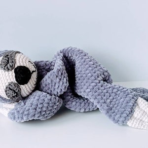May include: A gray crocheted sloth with white feet and a blanket. The sloth is lying on its side with its head resting on the blanket.