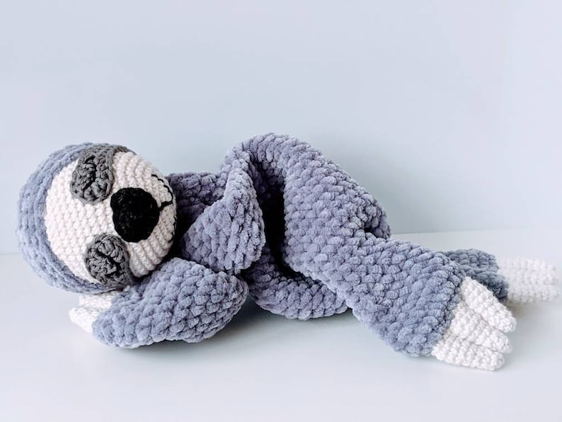 May include: A gray crocheted sloth with white feet and a blanket. The sloth is lying on its side with its head resting on the blanket.