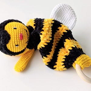 May include: A crocheted bee toy with a yellow and black striped body, white wings, and a wooden ring attached.