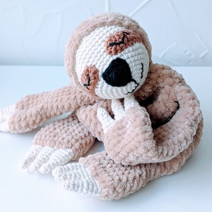 May include: A crocheted sloth toy with brown fur and white accents. The sloth has a sleepy expression with closed eyes and a small black nose.