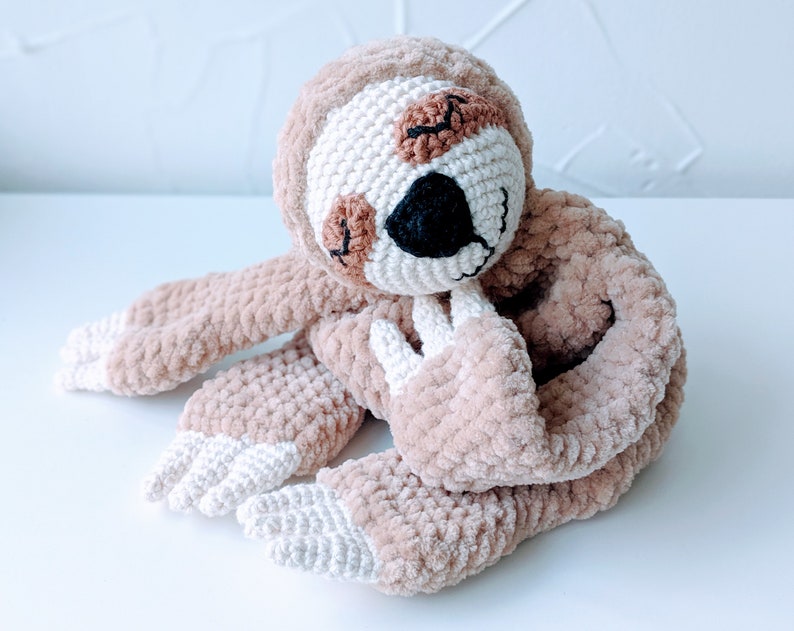 May include: A crocheted sloth toy with brown fur and white accents. The sloth has a sleepy expression with closed eyes and a small black nose.