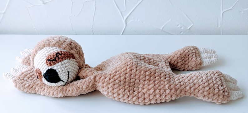 May include: A crocheted plush sloth toy in a light brown color with a white belly and brown eyes. The sloth is lying on its back with its arms and legs outstretched.