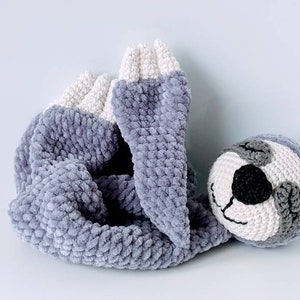 May include: A gray and white crocheted sloth toy with a black nose and eyes. The sloth is lying on its side with its arms and legs curled up.