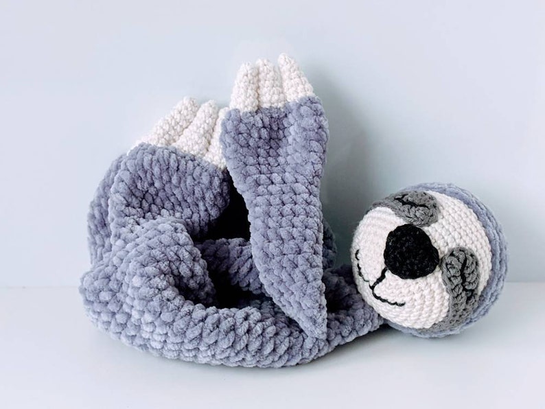 May include: A gray and white crocheted sloth toy with a black nose and eyes. The sloth is lying on its side with its arms and legs curled up.
