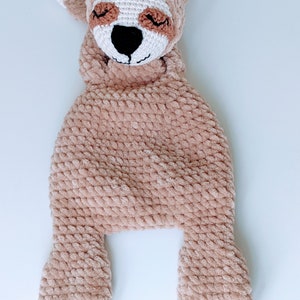 May include: A crocheted sloth toy with brown fur, white feet, and a brown face with closed eyes. The toy is lying on its back with its arms and legs outstretched.