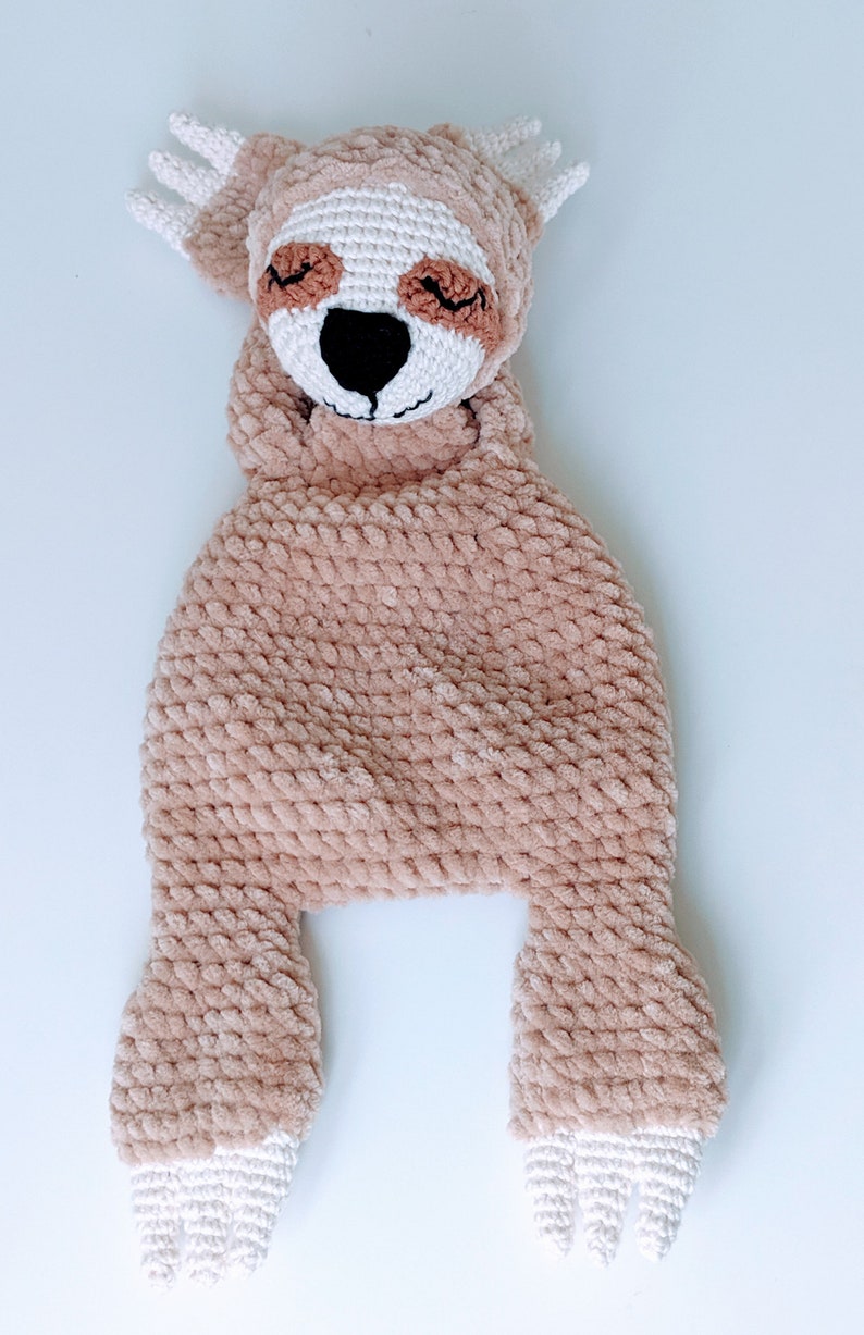 May include: A crocheted sloth toy with brown fur, white feet, and a brown face with closed eyes. The toy is lying on its back with its arms and legs outstretched.