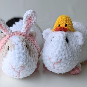May include: Two crocheted guinea pig plushies. One is wearing a pink bunny ear headband and the other is wearing a yellow duck hat.