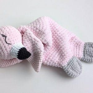 May include: Pink crocheted flamingo soft toy with grey feet. The flamingo is lying on its side with its head resting on its wing.