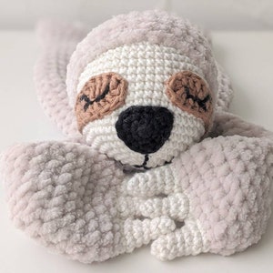 May include: A crocheted sloth toy with a light gray body, white face, and brown eyes. The sloth is lying on its stomach with its arms crossed in front of it.