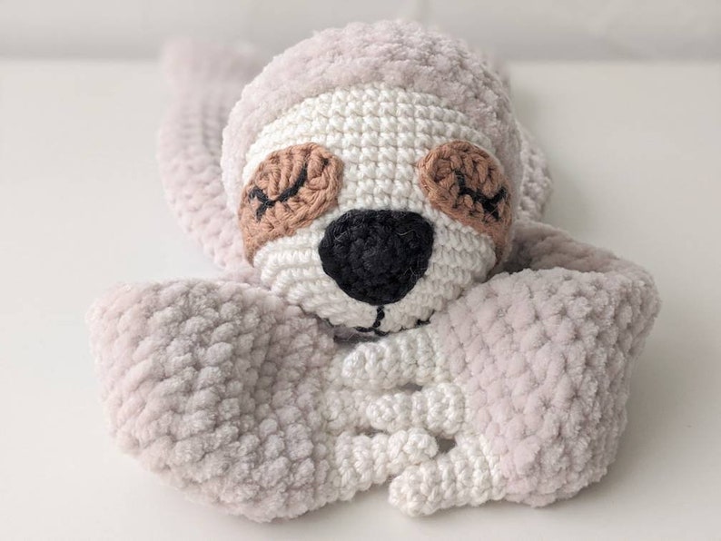 May include: A crocheted sloth toy with a light gray body, white face, and brown eyes. The sloth is lying on its stomach with its arms crossed in front of it.