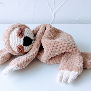 May include: A crocheted sloth plush toy with a brown and white color scheme. The sloth is lying on its side with its arms and legs outstretched. The sloth has a white blanket wrapped around it.