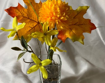 May include: A small glass vase filled with artificial flowers. The vase is clear and square shaped. The flowers are yellow and orange and include a large yellow chrysanthemum, yellow forsythia, and a single orange maple leaf.