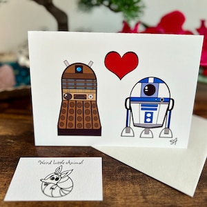 May include: A white card with a red heart, a brown and beige robot, and a blue and white robot. The card says "Weird Little Animal" at the bottom.