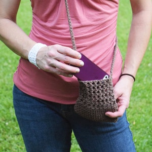 May include: A woman wearing a pink tank top and blue jeans is holding a brown crocheted bag with a purple phone inside.