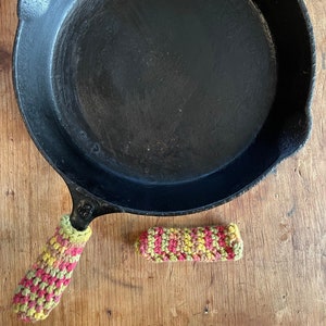 May include: A black cast iron skillet with a crocheted handle cover and a separate handle cover. The handle covers are yellow, pink, and green. The skillet is on a wooden surface.