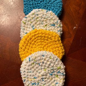 May include: Four crocheted dishcloths in a stack. The top dishcloth is white with green and blue flecks. The second dishcloth is yellow. The third dishcloth is white with green and blue flecks. The bottom dishcloth is blue.