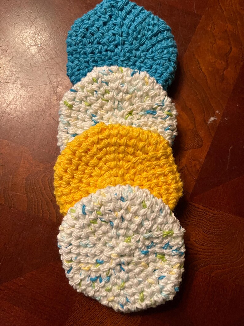 May include: Four crocheted dishcloths in a stack. The top dishcloth is white with green and blue flecks. The second dishcloth is yellow. The third dishcloth is white with green and blue flecks. The bottom dishcloth is blue.