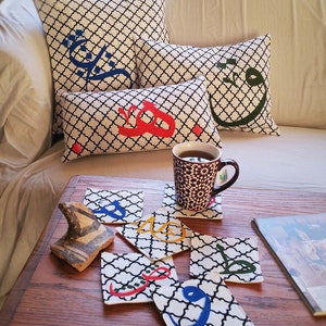 May include: A set of six black and white fabric coasters with colorful Arabic calligraphy. The coasters are on a wooden table with a mug of tea and a decorative ceramic object.