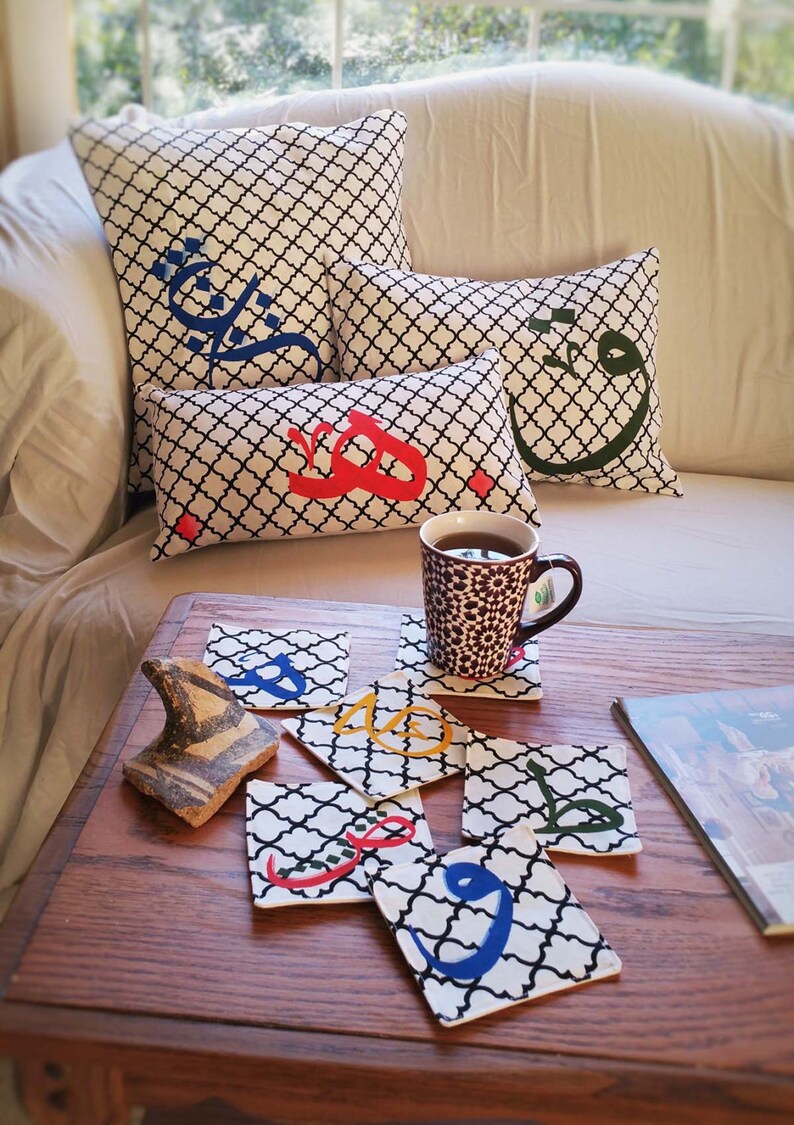 May include: A set of six black and white fabric coasters with colorful Arabic calligraphy. The coasters are on a wooden table with a mug of tea and a decorative ceramic object.
