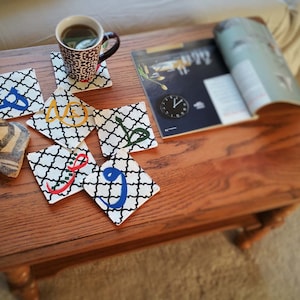 May include: A set of six square coasters with a black and white geometric pattern and colorful Arabic calligraphy. The coasters are arranged on a wooden table with a magazine and a cup of coffee.