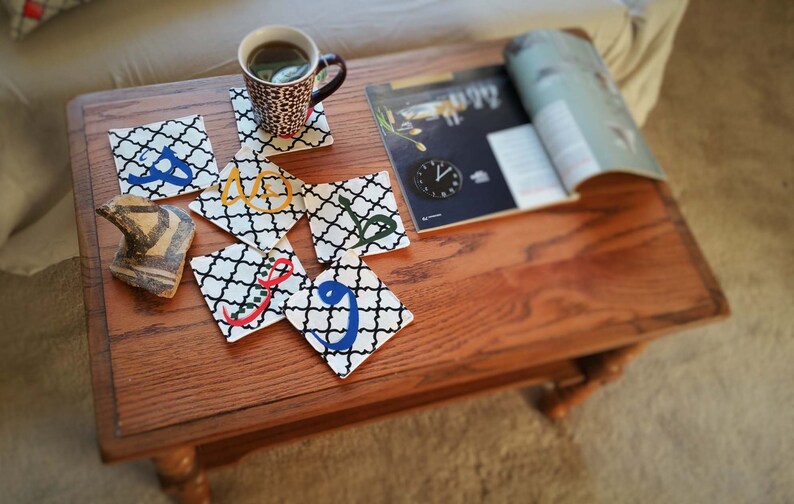 May include: A set of six square coasters with a black and white geometric pattern and colorful Arabic calligraphy. The coasters are arranged on a wooden table with a magazine and a cup of coffee.