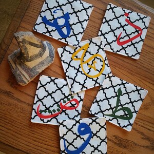 May include: Six square fabric coasters with black and white patterned designs. Each coaster features a different Arabic letter in a bright color, including red, blue, green, and yellow.