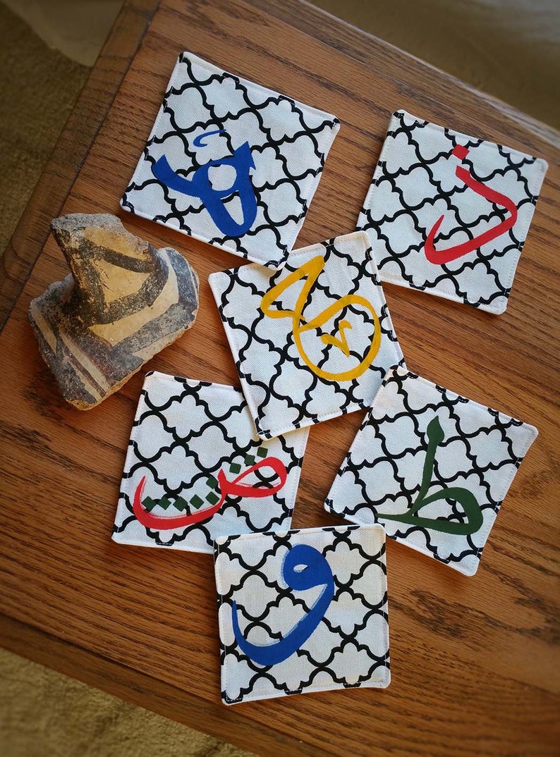May include: Six square fabric coasters with black and white patterned designs. Each coaster features a different Arabic letter in a bright color, including red, blue, green, and yellow.