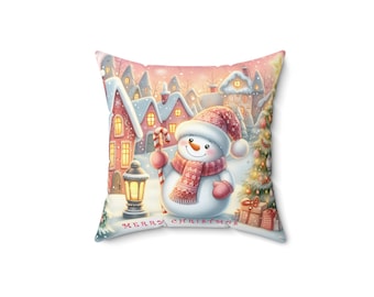 Op de afbeelding: Vierkant decoratief kussen met een grillige winterscène. Een sneeuwpop met een rode hoed en sjaal houdt een zuurstok vast voor besneeuwde huizen. De tekst "MERRY CHRISTMAS" staat onderaan.