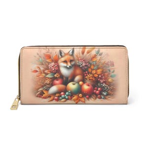 May include: A beige wallet with a gold zip and a colourful illustration of a fox surrounded by autumn foliage, apples, and berries. The fox has orange fur and a white chest. The wallet is rectangular.