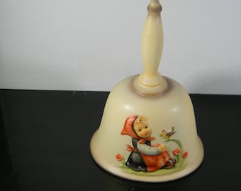 May include: A porcelain bell with a white and brown glaze. The bell features a painted scene of a girl in a red and black dress with a bird perched on a branch. The girl is looking at the bird and smiling.