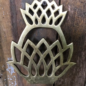 May include: A brass pineapple-shaped trivet with a decorative design. The trivet is attached to a wooden surface with a screw.